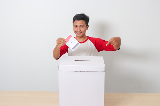 Portrait of excited Asian man inserting and putting the voting paper into the ballot box. General elections or Pemilu for the president and government of Indonesia. Isolated image on white background