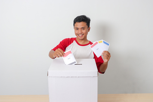 Portrait of excited Asian man inserting and putting the voting paper into the ballot box. General elections or Pemilu for the president and government of Indonesia. Isolated image on white background