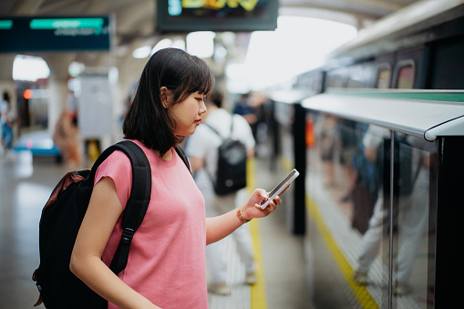 An Asian Chinese woman using smartphone while waiting for MRT train at station platform