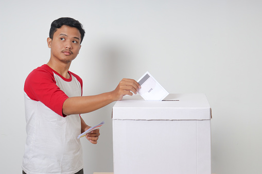 Portrait of excited Asian man inserting and putting the voting paper into the ballot box. General elections or Pemilu for the president and government of Indonesia. Isolated image on white background