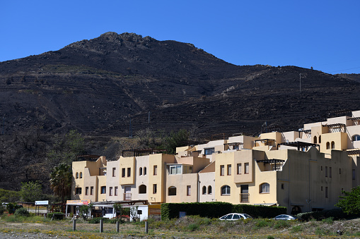 In the middle of the photo are houses, in the distance is a hill which has been devasted by a wildfire. The hill was growing vines which were destroyed in the fire.

The photo was taken at the Plage de Peyrefite, Peyrenees Orientales, France, Mediterranean near border with Spain. It was taken in May 2023.