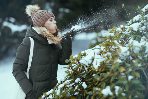 modern middle aged woman in green coat and brown hat outdoors in the city park in winter with mittens and beanie hat blowing snow near snowy branches.