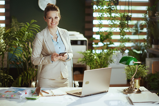 Portrait of happy modern small business owner woman in a light business suit in modern green office with laptop using smartphone applications.