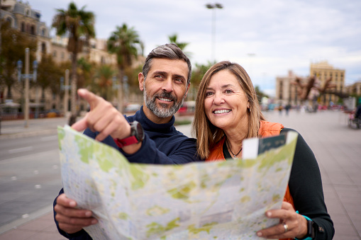 Middle-aged Caucasian couple with travel map in hands looking and pointing places of tourist interest. Husband and wife on street of city enjoying vacation happy and smiling. Adult tourism people