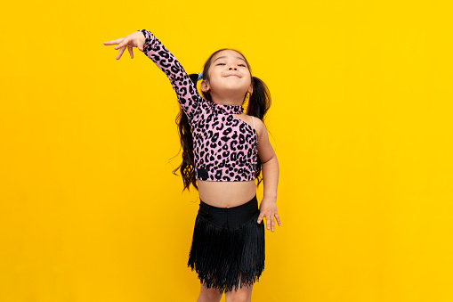 little asian girl in dance outfit dances chachacha on yellow isolated background, korean child dancer trains dance and stands in dance pose