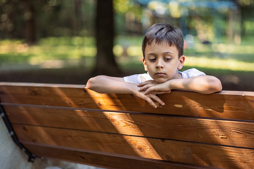 Little boy feeling sad while sitting on bench in the park