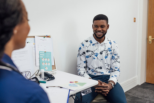 A young man visiting a medical clinic in Newcastle upon Tyne, England for a check up. He is sitting in a doctor's office and talking to the nurse about his medical condition while smiling and listening to the advice she's giving. The main focus is the male patient.