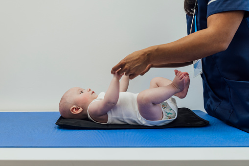 An unrecognisable mid adult nurse carrying out a medical exam on a baby boy in a medical clinic in Newcastle upon Tyne, England. The baby is lying on a table and baby changing mat in his vest while holding hands with the nurse and looking at her.