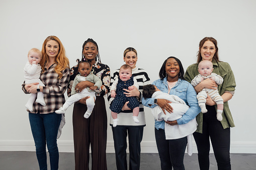 A portrait of a group of young women, holding their babies in their arms and standing in front of a white wall in a studio in Newcastle upon Tyne, England. They are all standing side by side, looking at the camera and smiling while most of the babies look curiously towards the camera and one is fast asleep.