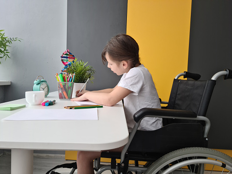 A focused young girl sits in a wheelchair at a white table, diligently drawing with colored pencils in a brightly lit room