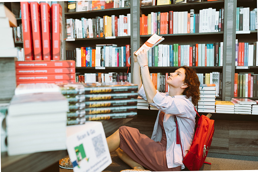 attractive girl student with brown curly hair, sitting on floor library, bookstore, holding book in hands, backpack on shoulders