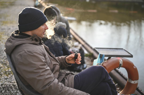 jovem fumando cigarro eletrônico na margem do rio. - quit lake - fotografias e filmes do acervo