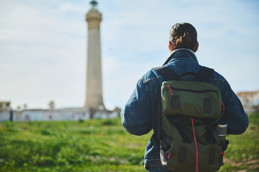 Young backpacked traveler hiker adventurer tourist man standing back to the camera and looking at a lighthouse background on a foggy day. People. Tour tourism. Travel and active lifestyle