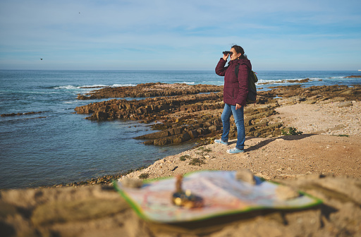 Young adult tourist traveler woman with a backpack, looking in binoculars standing on top of a cliff, enjoying a beautiful view of the Atlantic ocean. Blurred compass and geographic map on foreground.