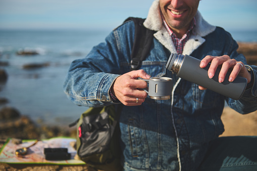 Happy smiling adventurer man in casual denim, pouring hot drink from a thermos flask into a stainless steel mug, sitting on a cliff by sea. Recreation. Active lifestyle. People and travel concept