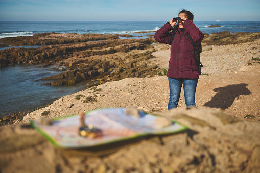 Young adult traveler woman adventurer looking into the distance through binoculars, standing on the top of a cliff by sea. People and active tourism concept. Copy advertising space