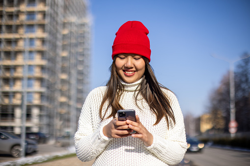 Young woman wearing sweater and red knit hat using smartphone in city during day