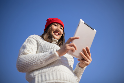 Young woman wearing sweater and red knit hat, using digital tablet in city during day, low angle view, clear sky, copy space