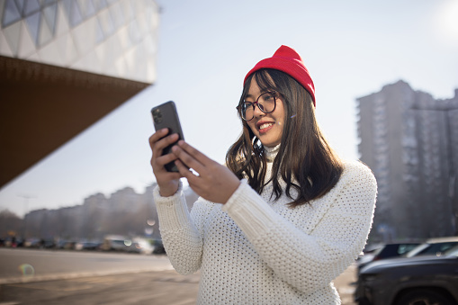 Young woman wearing sweater and red knit hat, taking a selfie in city during day
