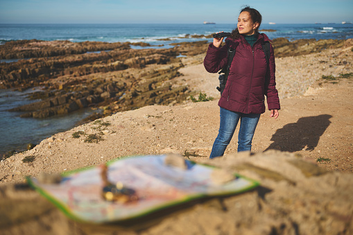 Active female adventurer traveler looking into the distance through binoculars, standing on the rock by sea. Blurred foreground with a navigational vintage old compass and map. Tourism. Travel concept