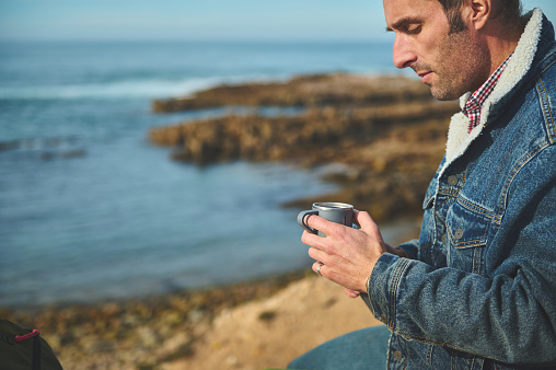 Close-up portrait of Caucasian relaxed serene man traveler holding a mug with hot drink in his hands, relaxing, sitting alone on a rock by sea, enjoying happy weekend on the nature. People. Lifestyle.