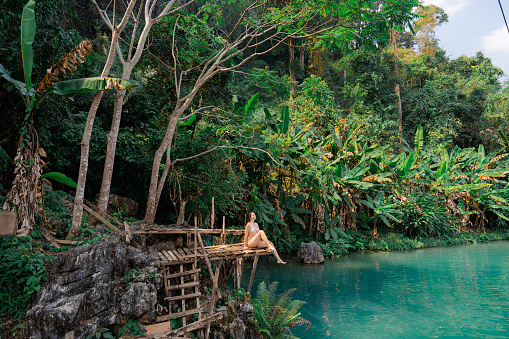 A woman sits peacefully on a rustic wooden dock by a tranquil, turquoise jungle river surrounded by lush tropical foliage.