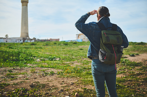 Rear view of a traveler hiker man with backpack, standing back to the camera and looking into the distance, standing over a lighthouse background on a foggy day. People. Travel and active lifestyle