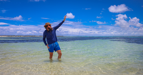 A man stands in the ocean off the coast of Mauritius, raising his arms in the air.