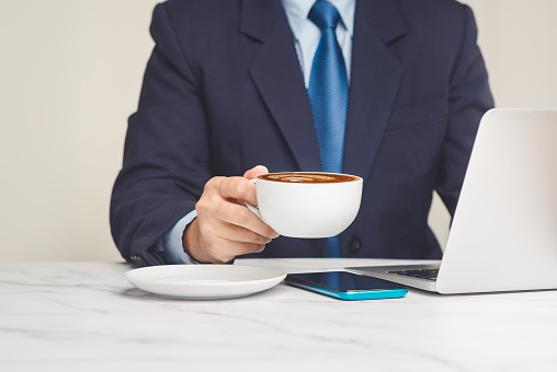 Businessman in a suit holding a cup of coffee while working at a desk in the office. Close-up photo. Beverage and relaxation concept