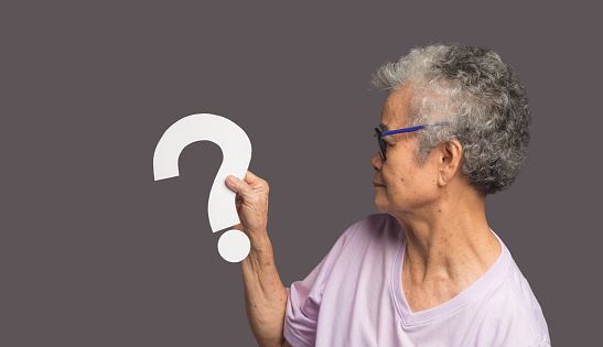 Question mark concept. An elderly Asian woman with short white hair holding a white question mark symbol with a smile while standing on a gray background. Space for text