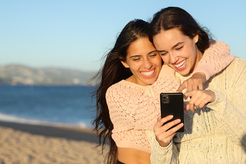 Two joyful friends checking phone on the beach