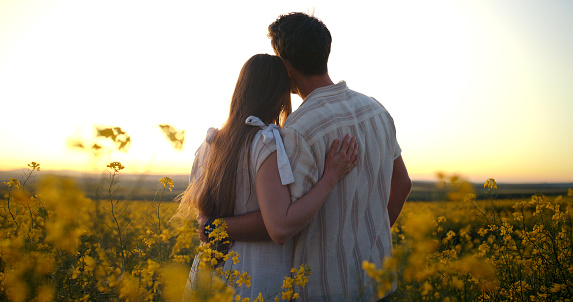 Travel, sunset and couple in canola field walking on vacation, holiday or weekend trip. Adventure, nature and back of man and woman on journey in outdoor park with yellow flowers in countryside.