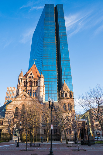 In the heart of Boston, as the early morning light bathes the city in a tranquil glow, the iconic Trinity Church stands in solemn reverence. Its historic Gothic architecture, a testament to the city's rich cultural heritage, presents a striking contrast against the backdrop of a modern skyscraper. The streets, usually bustling with activity, are uncharacteristically empty, accentuating the peaceful ambiance of the dawn.

The clear blue sky serves as a perfect canvas, highlighting the church's impressive façade and the contemporary glass and steel structure behind it. This juxtaposition of the old and the new is a vivid representation of Boston's unique urban landscape, where historic landmarks seamlessly blend with modern architectural marvels.

The serene setting is devoid of the usual city traffic, offering a rare moment of calm in the busy metropolitan area. The quietness around Trinity Church allows for an uninterrupted appreciation of its majestic elegance and the aesthetic harmony it creates with the towering building behind it.

This early morning scene captures not just the physical beauty of Boston's architecture but also the timeless essence of the city's evolving narrative. It's a moment where reverence for the past meets the sleek sophistication of the present, a perfect embodiment of urban harmony and architectural beauty in one of America's most historic cities.