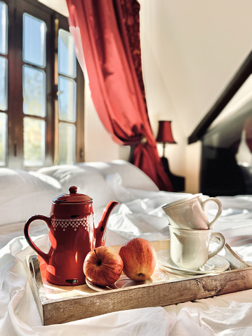 Two white faience cups, two ripe apples and a red coffee pot in blurry focus stand on a wooden tray on a white bed lit by bright morning light in vintage interior. Vertical image.