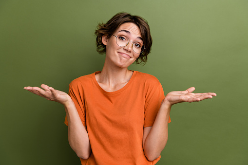 Portrait of indifferent woman with bob hairstyle wear stylish t-shirt in glasses shrugging shoulders isolated on khaki color background.