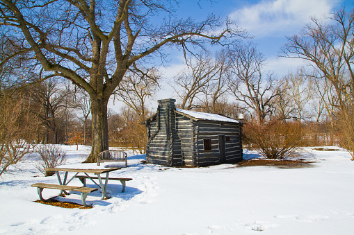 Tranquil winter scene featuring a historic log cabin with stone chimney, nestled in a snowy park setting in Fort Wayne, Indiana.