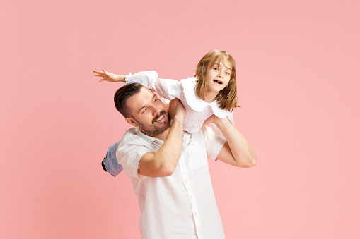 Man with playful smile carrying young girl on his shoulders, both simulating flight against pink pastel background. Concept of International Day of Happiness, childhood and parenthood. Ad
