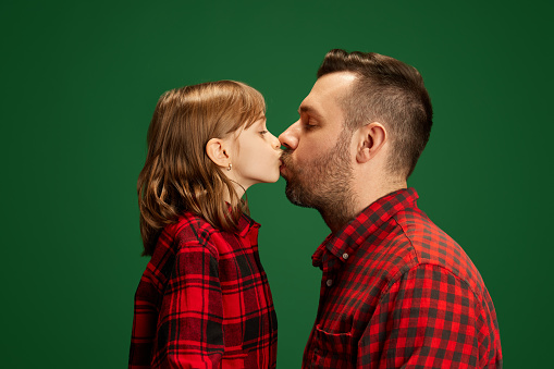 Side view portrait of father kissing his lovely daughter against green studio background. Concept of International Day of Happiness, childhood and parenthood, positive emotions. Ad