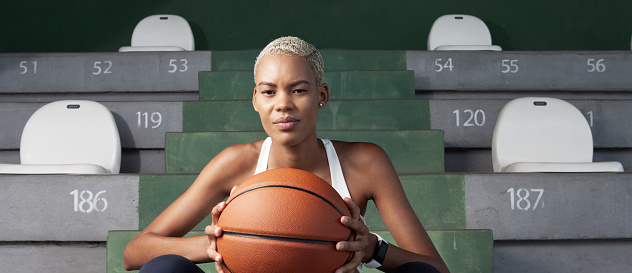 An African American female basket player sitting on the stadium stands, woman holding a basketball, embodying the concept of passion for the sport, game and physical training