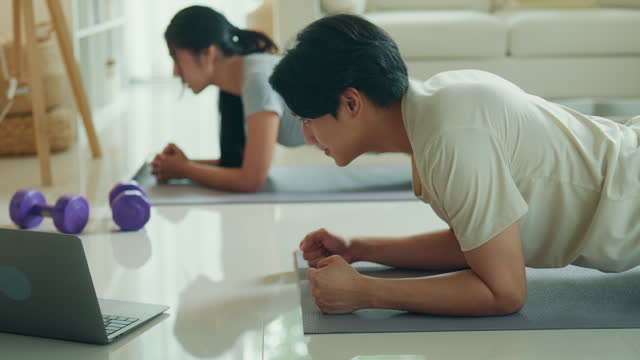 Cheerful young Asian couple is deeply concentrated on holding a plank position during a home workout session, following an online class. Enjoying home fitness workout.