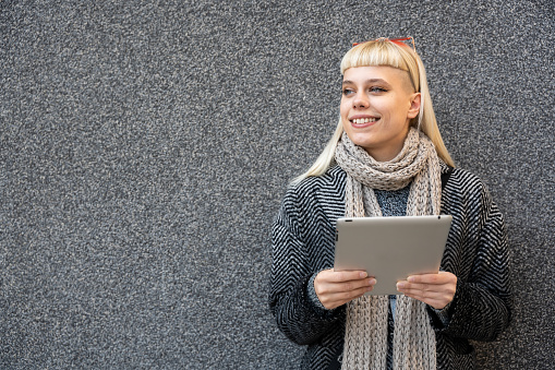 Happy hippie girl browsing a tablet working freelance and leaning on a wall of office building in the street. Hipster woman online work while waiting colleagues for staff meeting