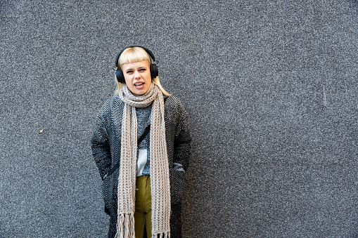 Young lovely hippie hipster woman in modern clothes wearing wireless headset listening music while leaning on the wall. Blonde female with cool attitude with headphones waiting on the street.