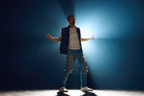 Casual dressed male singer with microphone intense performance under spotlight in darkness in dramatic smoke on stage. Concept f music and dance, lifestyle, festivals, concerts, self-expression.