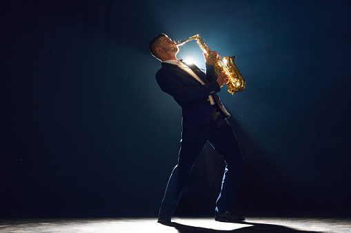 Jazz musician in suit virtuously playing saxophone stands with dramatic backlight behind him on dark stage. Concept of art, instrumental music, dance, culture, festivals and concerts.
