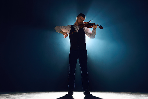 Violin player wearing formal outfit stands on stage, captured in moment of musical ecstasy, against dark background with light. Concept of instrumental music festivals and concerts, art, culture. Ad
