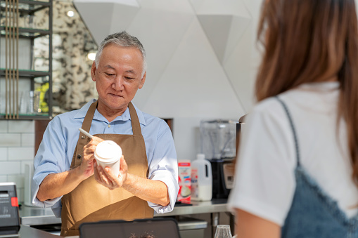 Coffee shop owner takes orders from happy customers. Dedicated shop owner stands behind the sleek bar counter, attentively taking orders from delighted female customers. Writes the customer's name on a coffee paper glass.