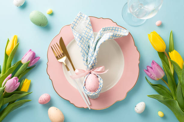 istock Bunny-themed Delight: Top-down view of an enchanting Easter table setting with bunny ear napkin, plate, cutlery, wine glass, tulips, and colorful eggs. Pastel blue background 1975265313