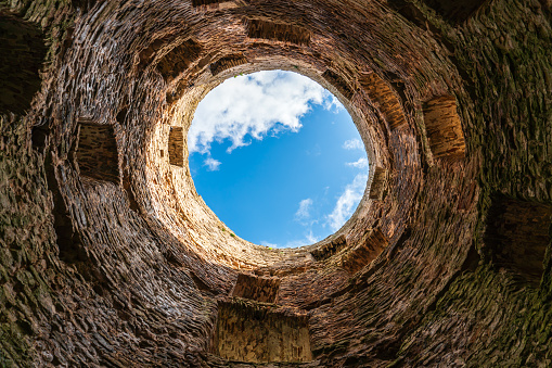 View of the sky with clouds from an old round medieval stone tower with powerful walls and narrow loopholes, the structure of natural stone, moss on the walls, cement masonry, details of fortress architecture, interesting background, history and culture.
