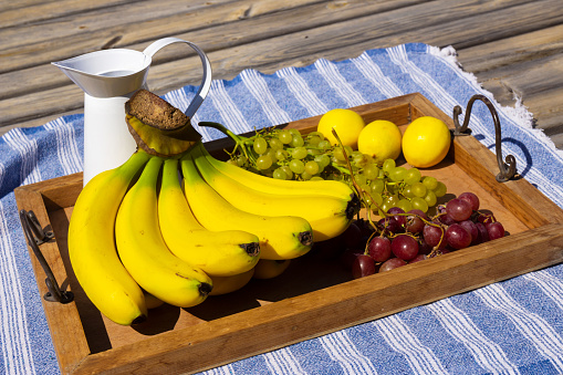 Variety of fresh ripe organic fruits on the wooden tray. Banana, grape and lemons on the blue tablecloth background on sunny day. Still life photo. Selective focus. Blurred background.