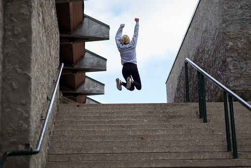 Blonde young woman with her back to the camera is jumping for joy and in triumph as she is reaching her fitness and health goals by running to the top of some steep stairs in the city. She is wearing a grey long sleeve and casual exercise clothing.